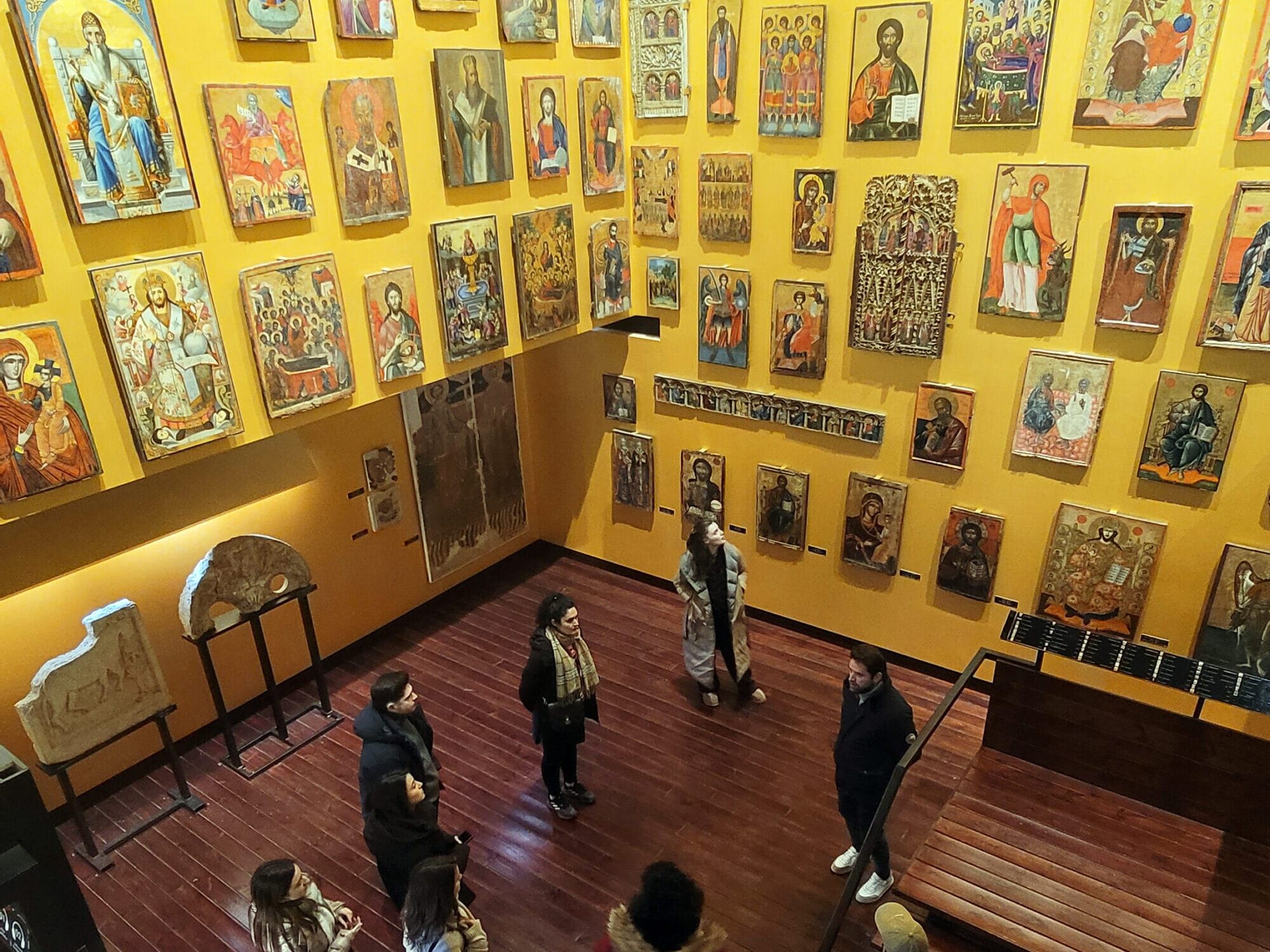 A group of visitors inside the National Museum of Medieval Art, standing on a wooden floor, surrounded by walls filled with religious icons.