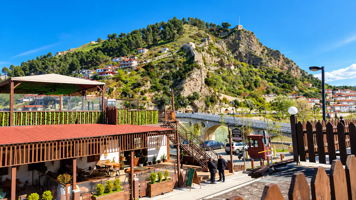 Scenic view of a traditional restaurant terrace near Gorica Bridge in Berat, Albania, with green hills and historic hillside houses.