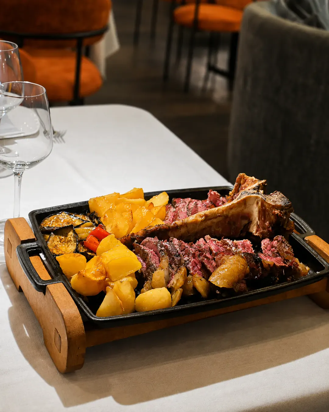 Biftek tomahawk i pjekur në skarë dhe i prerë në feta, i servirur me patate të pjekura dhe perime në një pjatancë të nxehtë në Idromeno Restaurant.