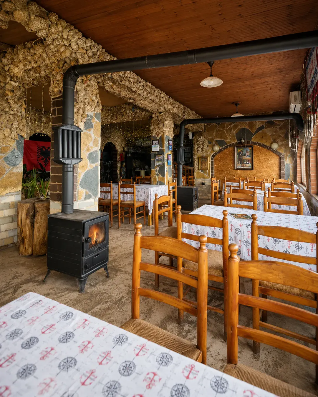 Ambient rustik i brendshëm i Taverna Shkodrane, me tavolina dhe karrige druri, mure guri, dekorime tradicionale dhe soba me dru në Shkodër, Shqipëri.