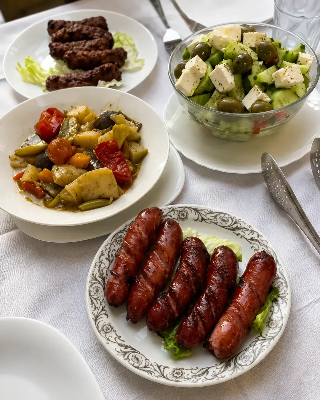 Salçiçe dhe qofte në skarë, të servirura me perime të përziera dhe sallatë të freskët me djathë të bardhë, kastravecë dhe ullinj.
