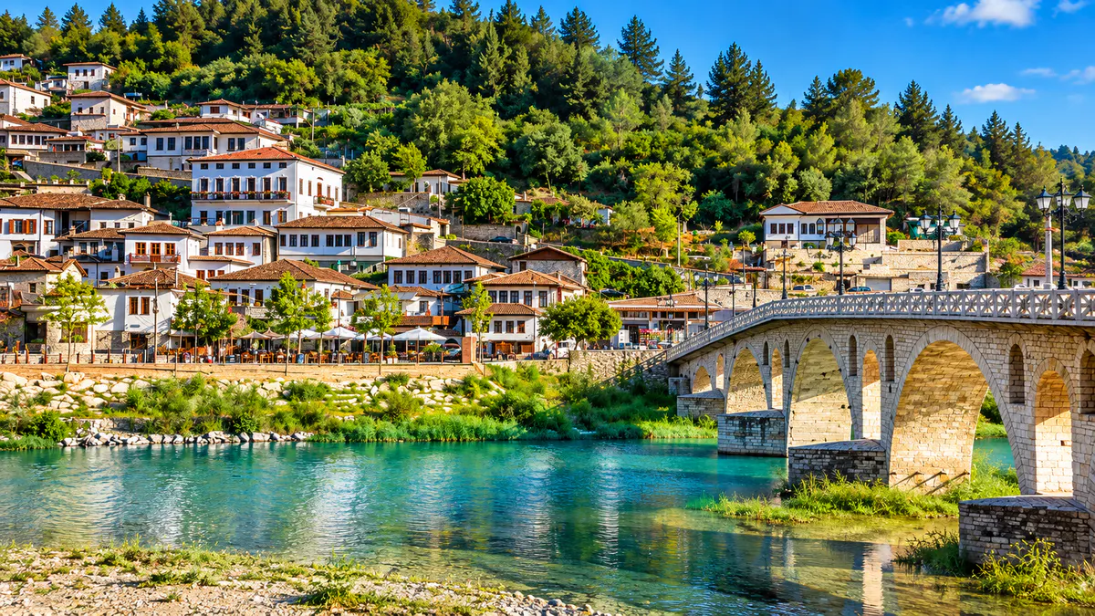 Ura historike e Goricës mbi lumin Osum në Berat, Shqipëri, me shtëpi tradicionale dhe kodra të gjelbra.