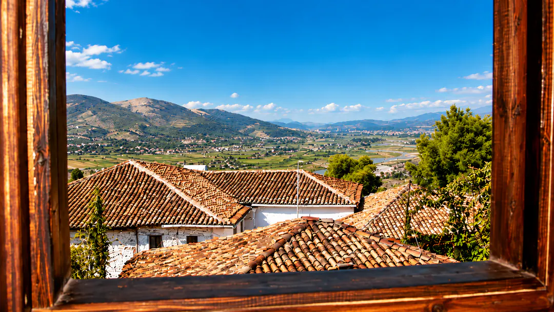 Panoramic view from Hotel Klea inside Berat Castle, featuring traditional red-tiled rooftops, green valleys, surrounding mountains and a clear blue sky.