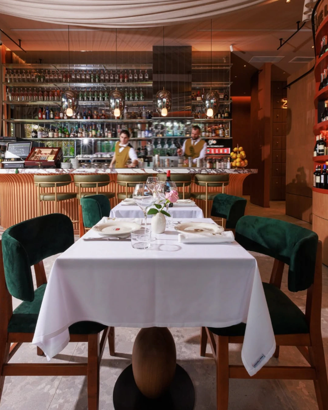 Elegant dining area at ZABÒ Garden in Tirana, featuring green velvet chairs, white tablecloths, and a stylish bar in the background.