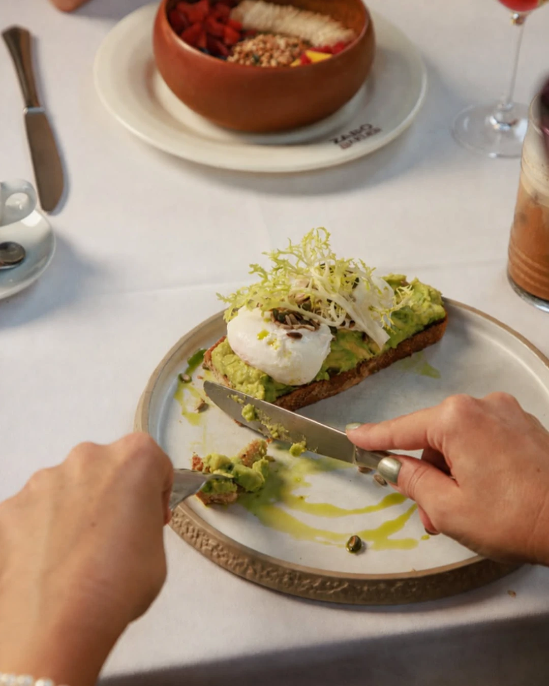 Close-up of a guest cutting avocado toast topped with a poached egg and fresh greens during brunch at ZABÒ Garden in Tirana.